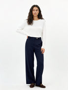 A woman modeling a white long-sleeve crewneck top and navy blue trousers, paired with dark brown loafers, against a plain white studio background.