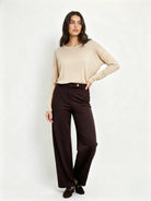 A woman modeling a beige long-sleeve knit top and dark brown wide-leg trousers against a plain light gray studio background.
