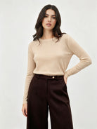 A woman modeling a beige long-sleeve crewneck knit top paired with dark brown wide-leg trousers, photographed against a clean light gray studio background.