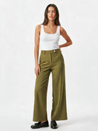 A model wearing olive green wide-leg trousers with a button waistband, styled with a white tank top and black loafers, against a plain white studio background.