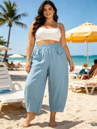 Woman in light blue pants and white top standing on a beach with palm trees and umbrellas in the background.