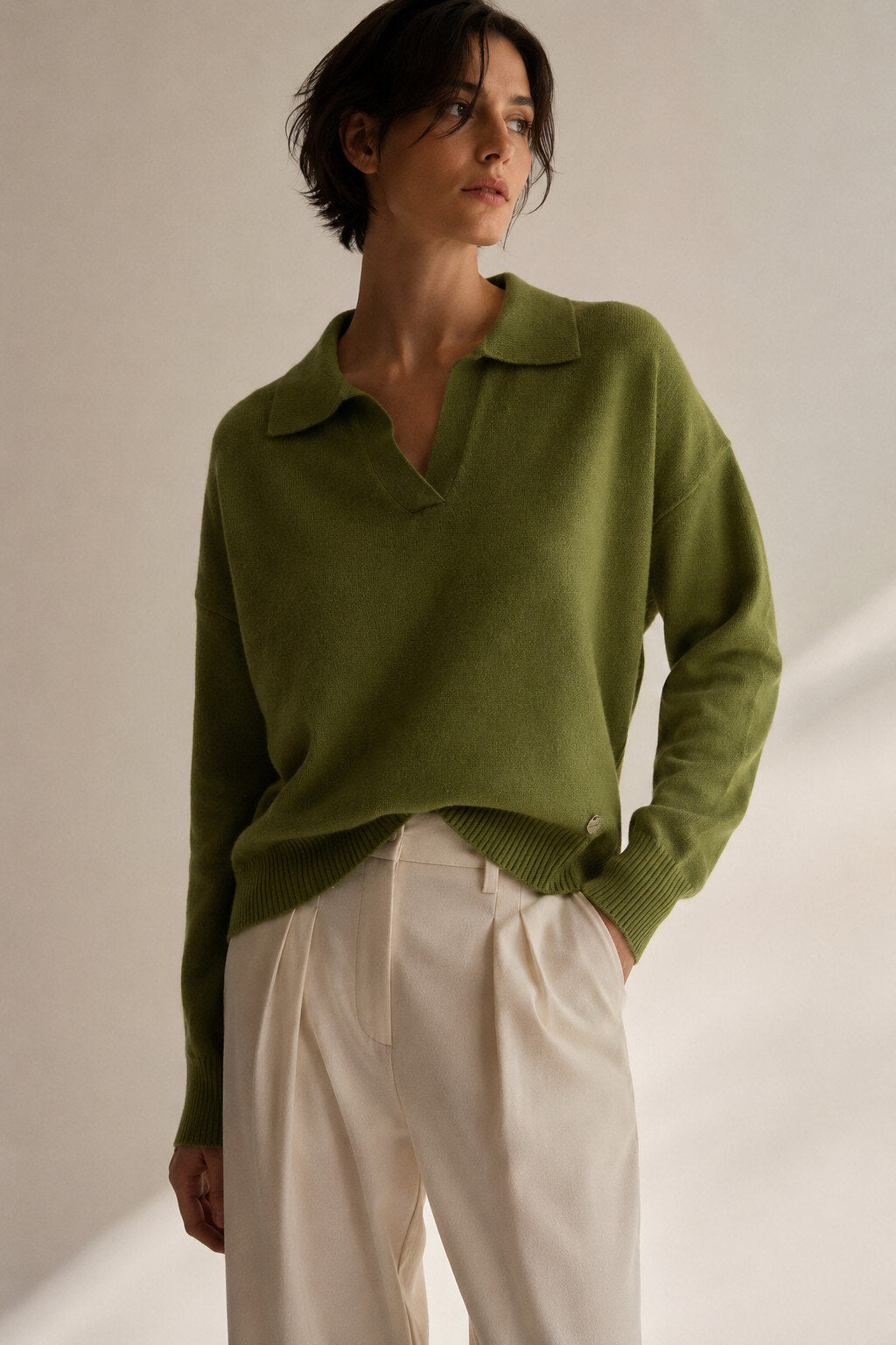 A woman wearing an olive green knit polo sweater and cream-colored trousers, standing against a warm beige background with soft shadows.