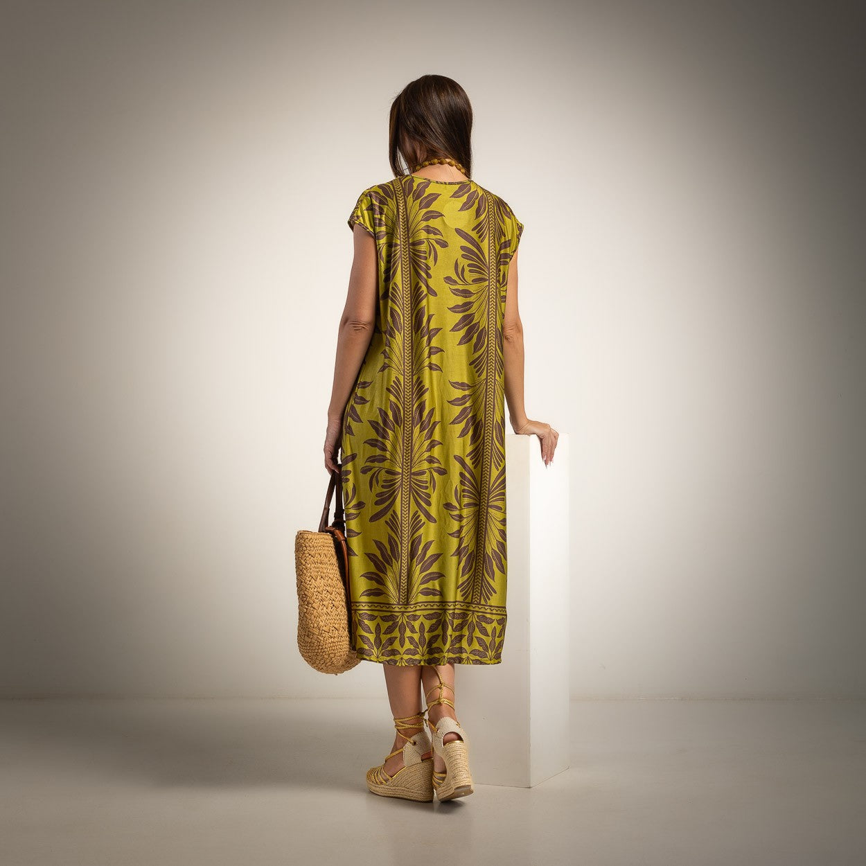 A woman stands with her back to the camera wearing a sleeveless olive green midi dress with a brown palm leaf pattern, holding a woven straw tote bag, set against a neutral studio background.