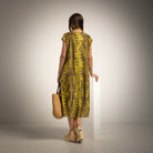 A woman stands with her back to the camera wearing a sleeveless olive green midi dress with a brown palm leaf pattern, holding a woven straw tote bag, set against a neutral studio background.