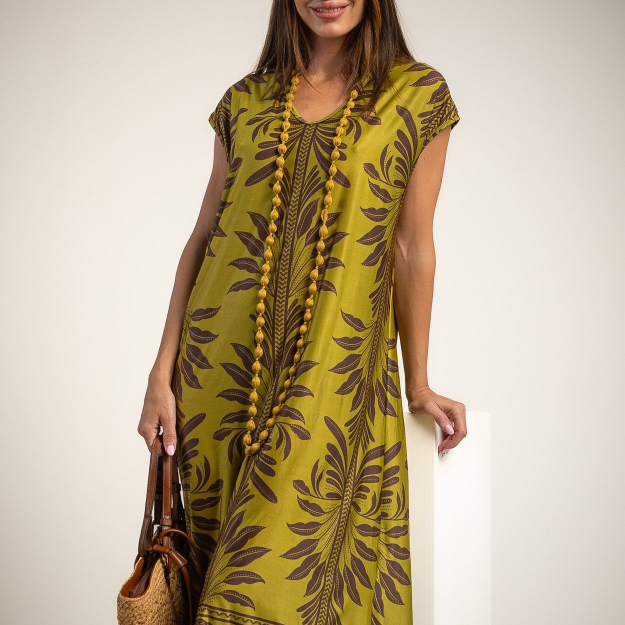 A woman wearing a short-sleeve kaftan dress in olive green with a dark brown botanical leaf pattern, accessorized with a long beaded necklace and holding a woven handbag.