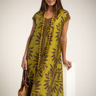 A woman wearing a short-sleeve kaftan dress in olive green with a dark brown botanical leaf pattern, accessorized with a long beaded necklace and holding a woven handbag.