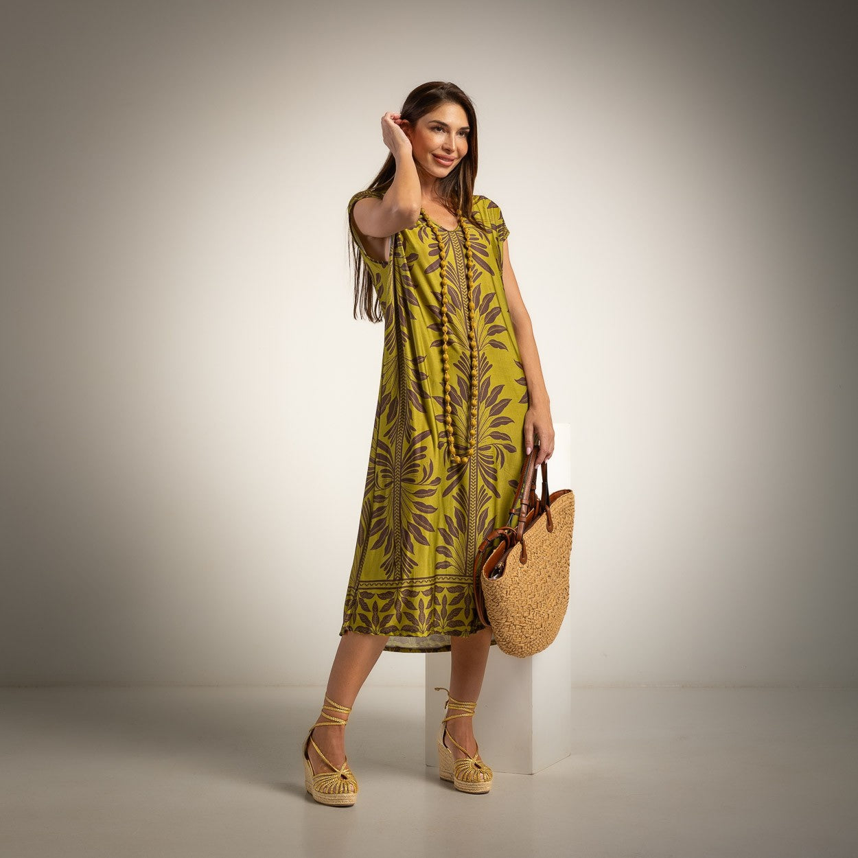 A woman models a sleeveless olive green midi dress with a brown botanical pattern, holding a woven straw tote bag and standing on a white pedestal against a neutral studio background.