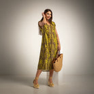 A woman models a sleeveless olive green midi dress with a brown botanical pattern, holding a woven straw tote bag and standing on a white pedestal against a neutral studio background.