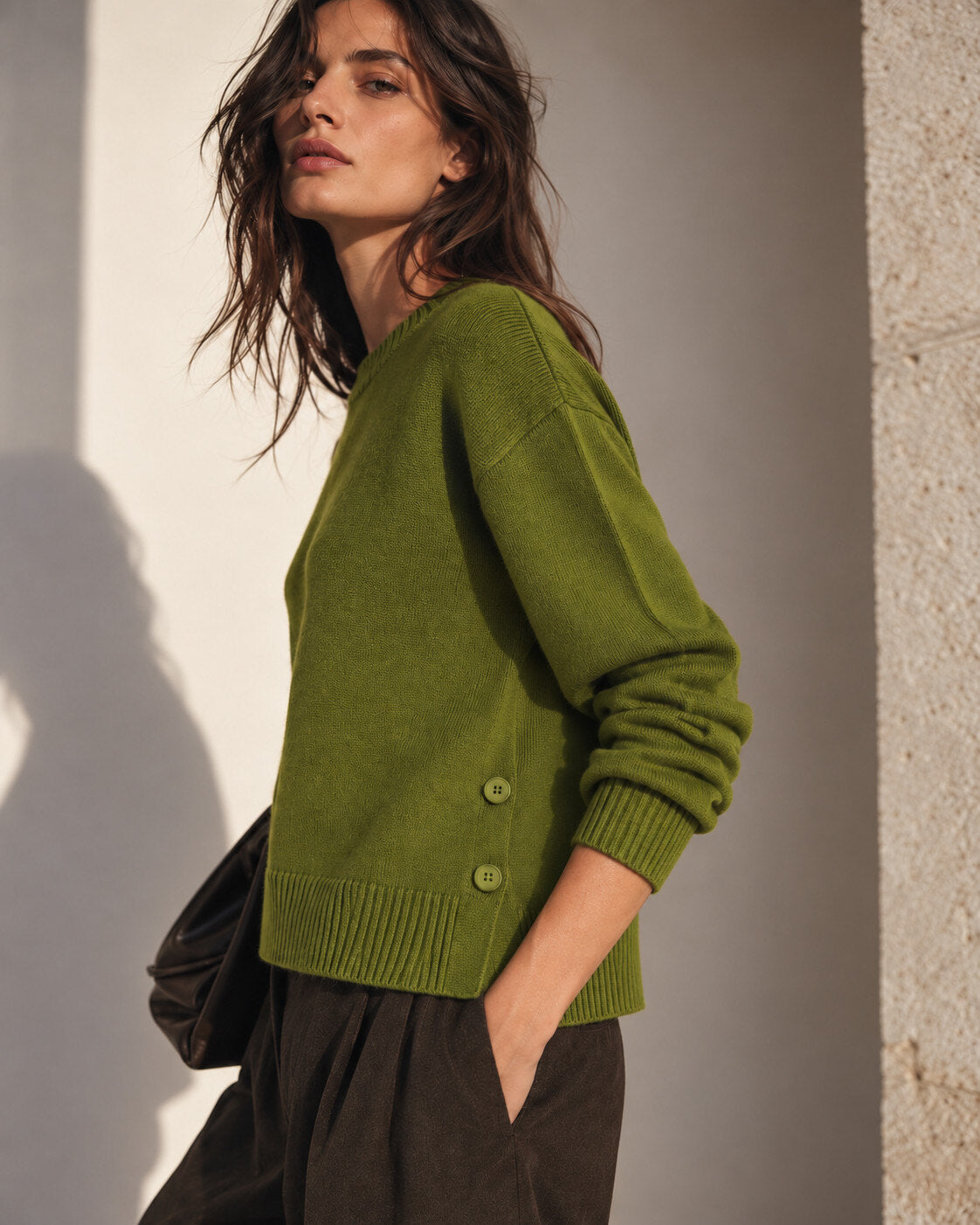 A woman wearing an olive green knit sweater with buttons on the side, paired with dark brown trousers, standing against a textured wall with warm sunlight and shadows.