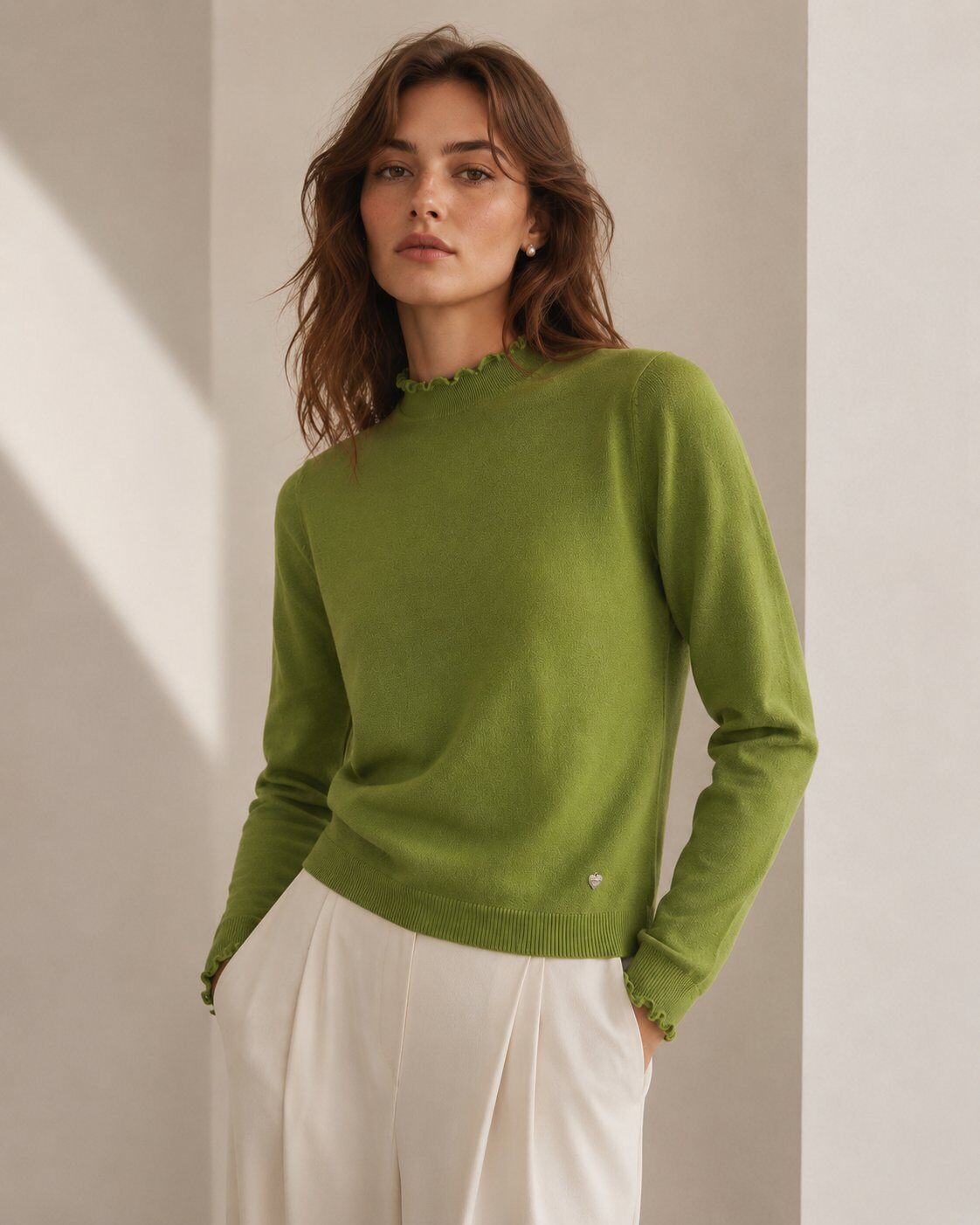 A woman wearing a light olive green knit sweater with a scalloped mock neck and white trousers, standing in a minimalist beige room with soft diagonal shadows.