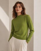 A woman wearing a light olive green knit sweater with a scalloped mock neck and white trousers, standing in a minimalist beige room with soft diagonal shadows.