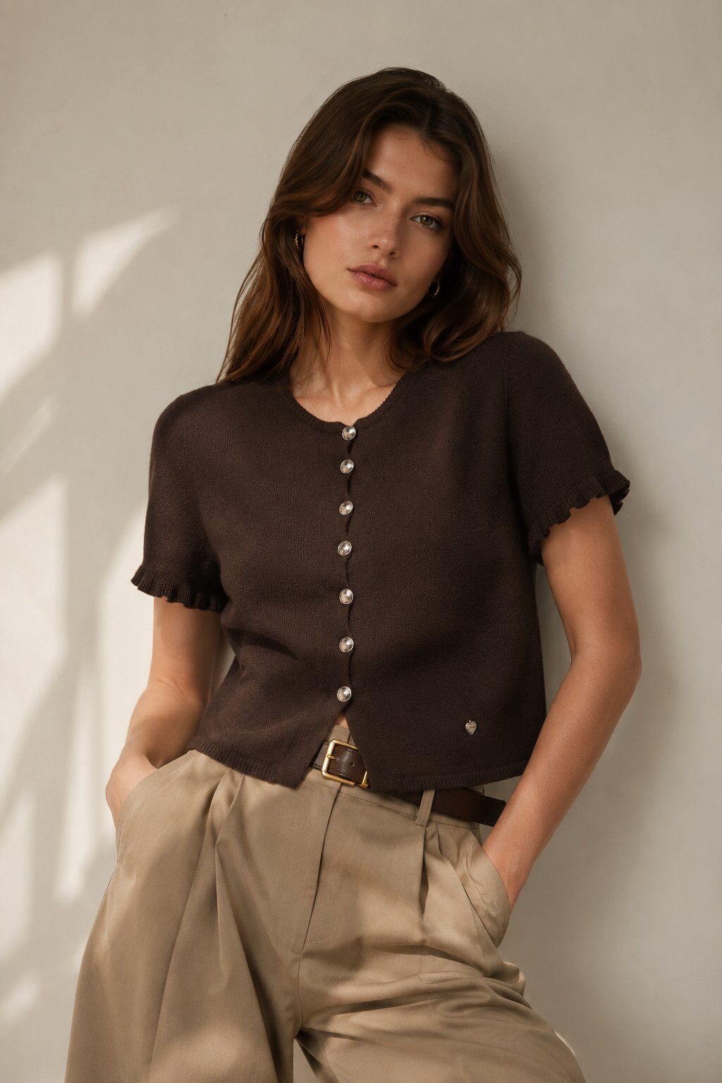 A woman wearing a dark brown short-sleeve knit top with scalloped edges and a button-down front, paired with beige trousers, leaning against a neutral wall.