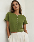 A woman wearing a cropped olive green knit top with short sleeves and a vertical row of pearl-like buttons down the front, paired with white trousers, standing against a plain off-white wall.