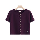 A short-sleeve knit top in deep plum purple, hanging on a wooden hanger against a plain white background. The top features a round neckline, a front button closure with buttons, and a small heart detail near the lower right hem.