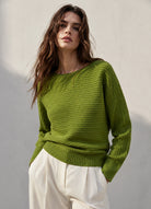 A woman wearing a textured olive green knit sweater and white trousers, standing against a neutral light grey wall.
