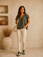 A woman stands in a warm, earthy-toned room wearing an olive green short-sleeve knit top, beige skinny pants, and dark sneakers, holding a beige shoulder bag.