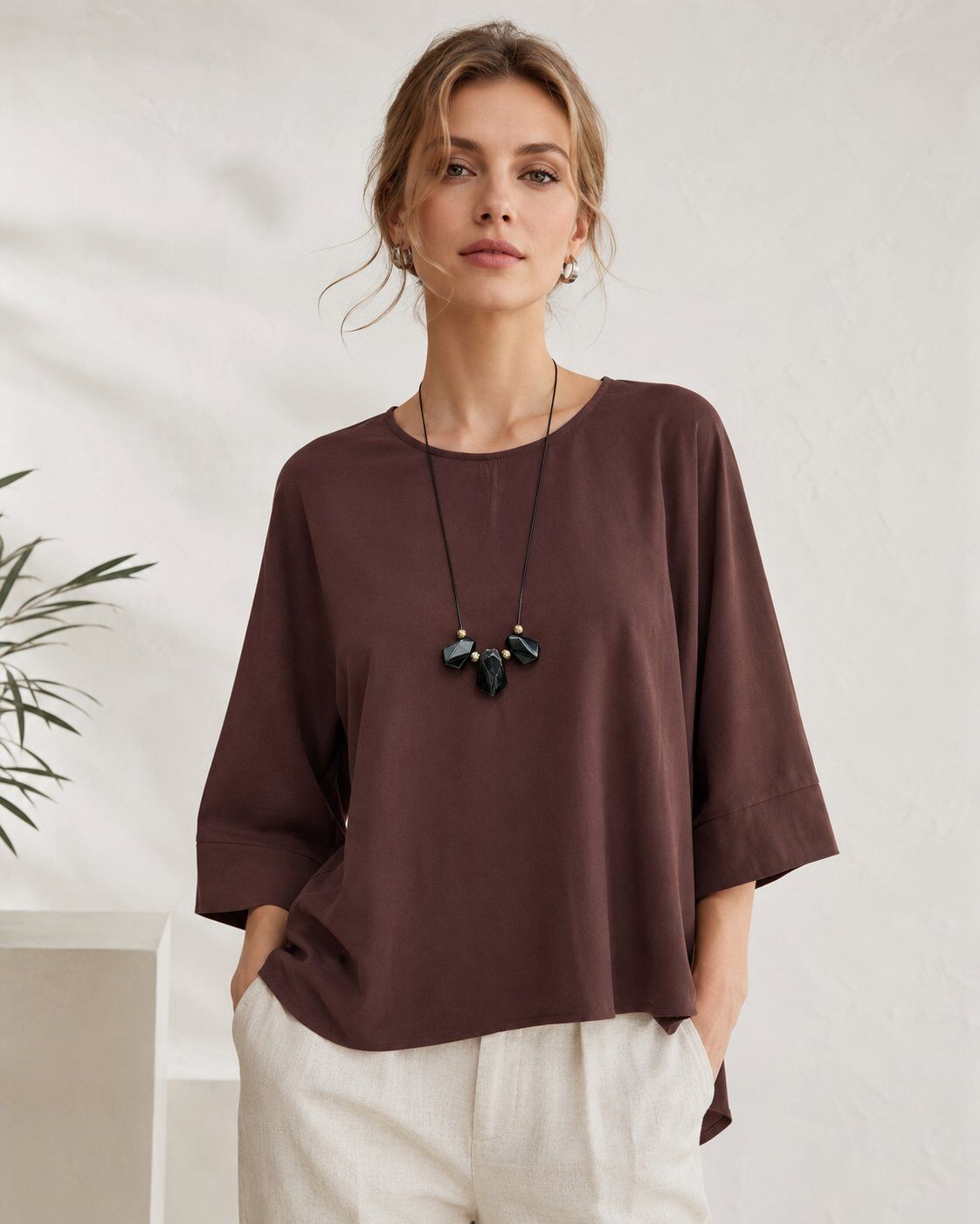 A woman wearing a loose-fitting, dark brown short-sleeve top with three-quarter sleeves, paired with off-white trousers and a black pendant necklace