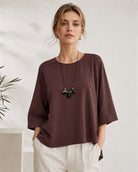 A woman wearing a loose-fitting, dark brown short-sleeve top with three-quarter sleeves, paired with off-white trousers and a black pendant necklace