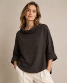 A woman wearing a dark brown cowl-neck poncho top with rolled three-quarter sleeves and white trousers, standing against a neutral beige studio background.