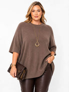 A woman modeling a brown knit poncho sweater with elbow-length sleeves, paired with dark brown faux-leather pants, a gold teardrop pendant necklace, and a brown crossbody bag, against a plain white background.