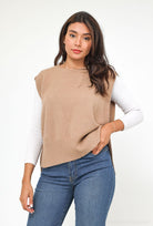 Woman wearing a beige sleeveless sweater over a white long-sleeve shirt with blue jeans on a white background