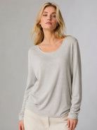 A woman modeling a light beige long-sleeve scoop-neck knit top, paired with cream pants, against a solid light gray studio background.
