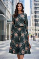 A woman stands on a city sidewalk wearing a dark green long-sleeve top and matching pleated A-line skirt in a green, pink, and beige plaid pattern. She is holding a brown leather handbag.