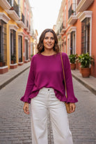A woman stands on a cobblestone street wearing a magenta long-sleeve top with ruffled cuffs and hem, paired with white wide-leg pants. She has a thin tan shoulder bag strap visible, with colorful colonial-style buildings in the background.