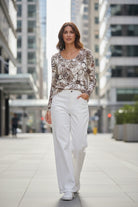A woman walking down a city street wearing a beige and brown floral long-sleeve top with white wide-leg trousers and white sneakers.