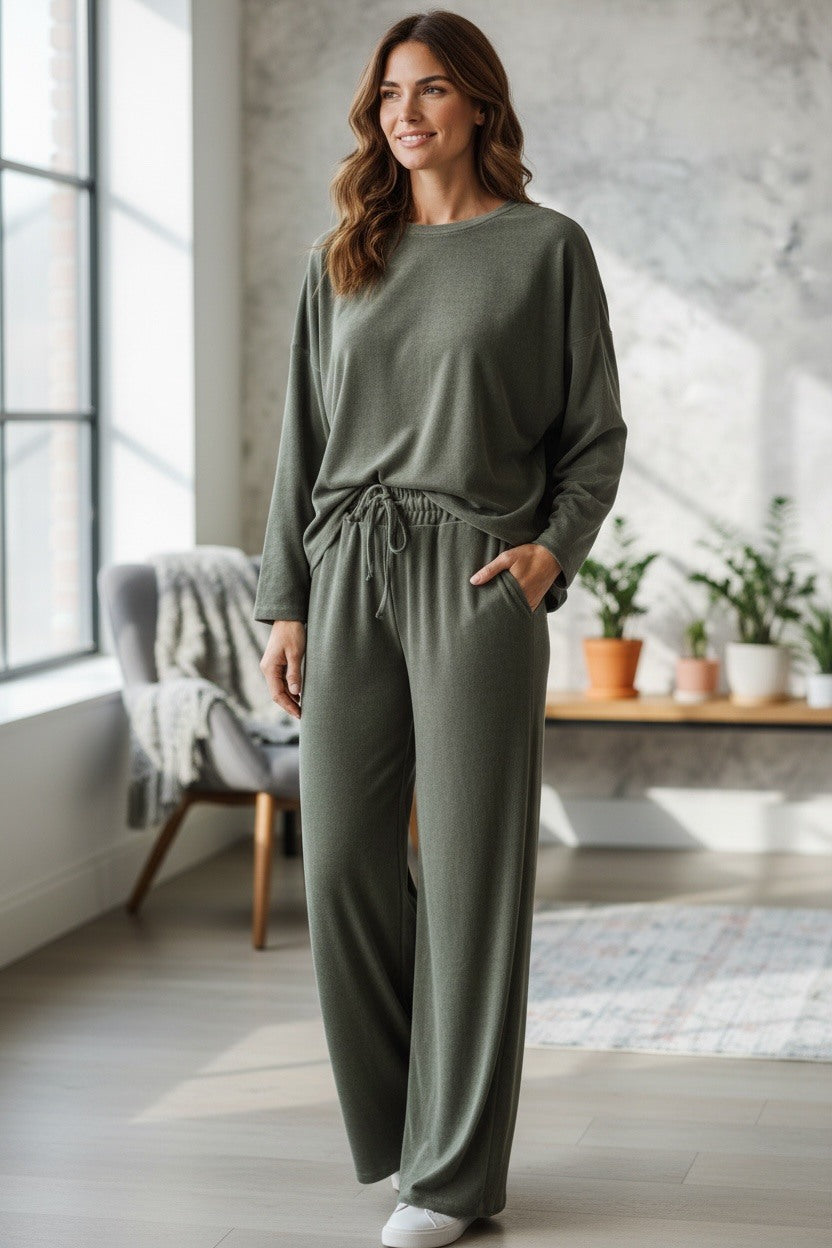 A woman modeling a matching two-piece lounge set in a bright, modern room. The set consists of a long-sleeve crewneck top and matching wide-leg drawstring pants in an khaki green color.