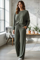 A woman modeling a matching two-piece lounge set in a bright, modern room. The set consists of a long-sleeve crewneck top and matching wide-leg drawstring pants in an khaki green color.