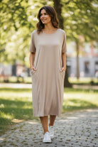 A woman wearing a short-sleeve beige midi dress with side pockets walks on a cobblestone path in a sunny park.