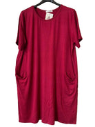 A short-sleeve crewneck dress in a burgundy colour, hanging on a black hanger against a textured gray wall. The dress features side pockets and a small white brand tag at the neckline.