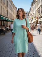 Woman in a light blue dress holding a coffee cup and a brown bag on a city street.