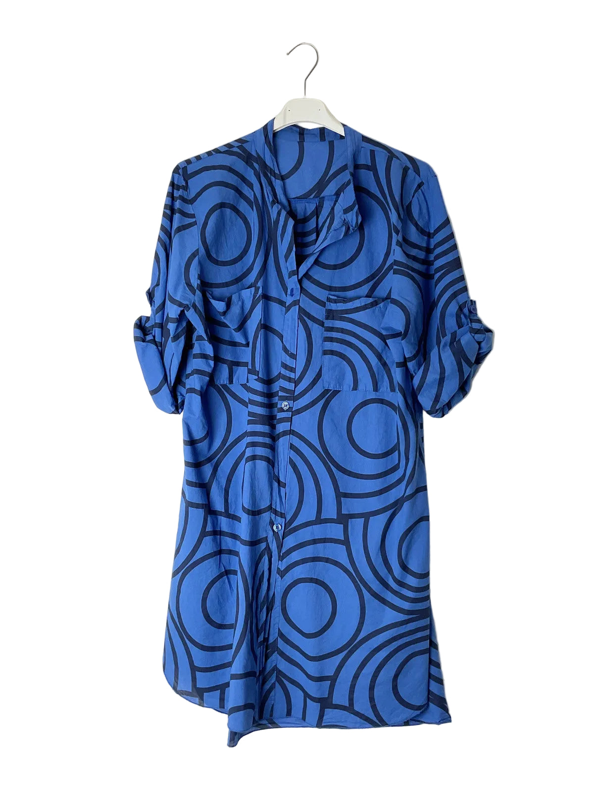 A blue button-up shirt dress with a black geometric circle pattern, hanging on a white hanger against a plain white wall.
