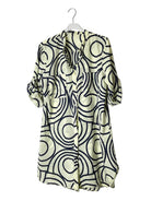A short-sleeve button-front shirt dress with a cream base and a bold black wavy geometric pattern, hanging on a white plastic hanger against a plain white wall.