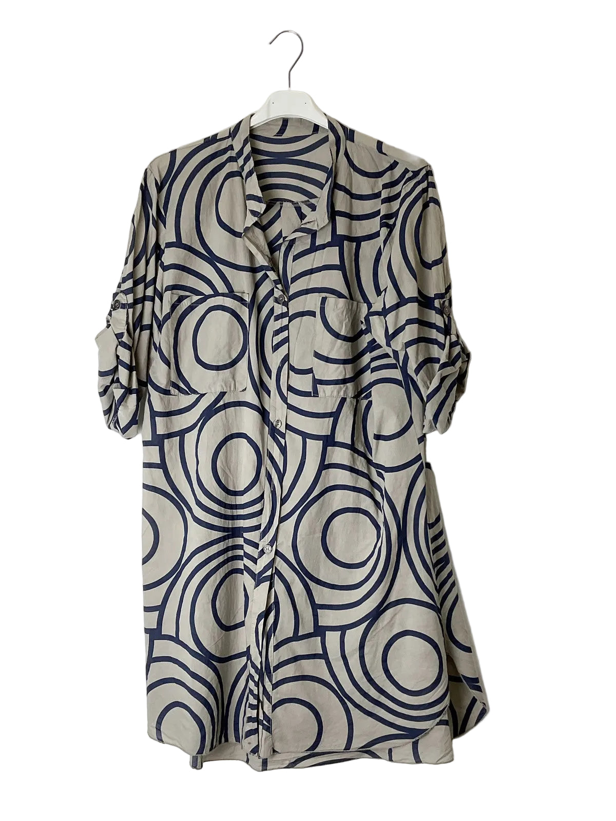 A long-sleeve button-up shirt dress with a beige base and a navy blue abstract circular pattern, hanging on a white hanger against a plain white wall.