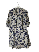A long-sleeve button-up shirt dress with a beige base and a navy blue abstract circular pattern, hanging on a white hanger against a plain white wall.