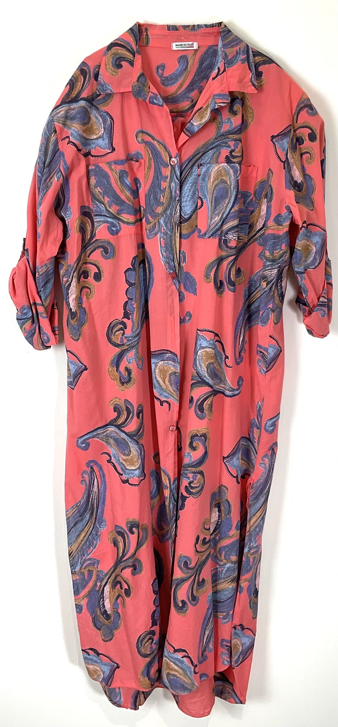 A coral pink long-sleeve button-up shirt dress with a blue, purple, and gold paisley pattern, shown hanging against a plain white background.