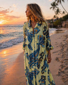 A woman stands on a beach at sunset wearing a long-sleeve button-front dress with a pale yellow base and a bold blue and dark brown ornamental print.