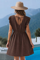 A woman viewed from behind wearing a brown textured mini dress with ruffled cap sleeves and a tie-back neckline, standing outdoors near a pool with mountains in the background.