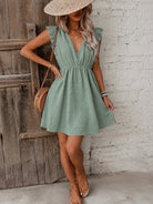 A woman wearing a light green textured mini dress with ruffle sleeves and a V-neckline, styled with a straw hat, a round tan crossbody bag, and tan heeled sandals, posing against a rustic wooden door and white brick wall.