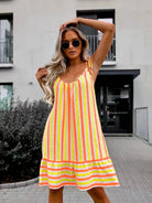 A woman wearing a sleeveless midi dress with vertical neon yellow, orange, and white stripes, styled with round sunglasses, posing outdoors in front of a modern building.