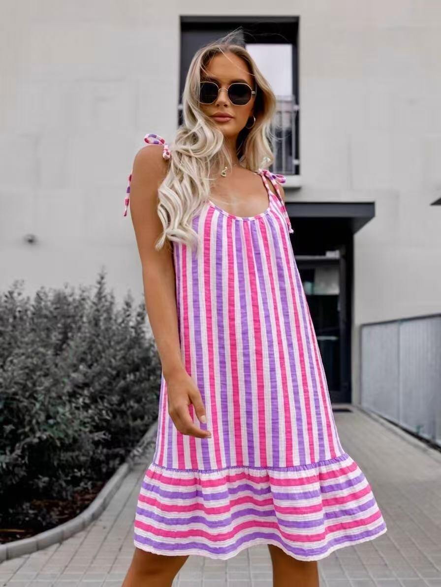 A woman wearing a pink, purple, and white vertically striped sleeveless dress with tie-shoulder straps and a ruffled hem, standing outdoors in front of a modern building.