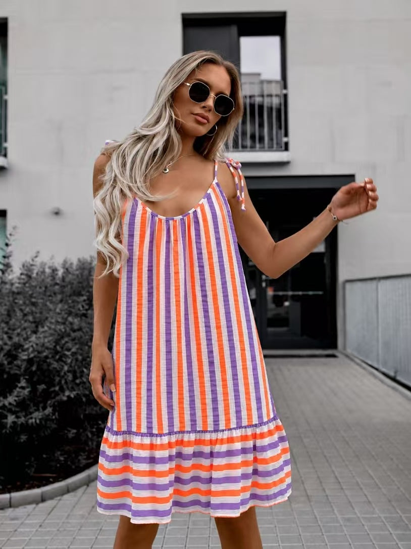 A woman wearing a sleeveless tiered dress with vertical orange, purple, and white stripes, styled with round sunglasses, standing outdoors in front of a modern building.