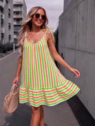 A woman wearing a sleeveless, tiered A-line dress with vertical lime green, orange, and white stripes, walking outdoors on a city street next to a concrete wall.