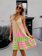 A woman wearing a sleeveless, tiered A-line dress with vertical pink and lime green stripes, styled with round sunglasses, standing outdoors on a paved walkway.