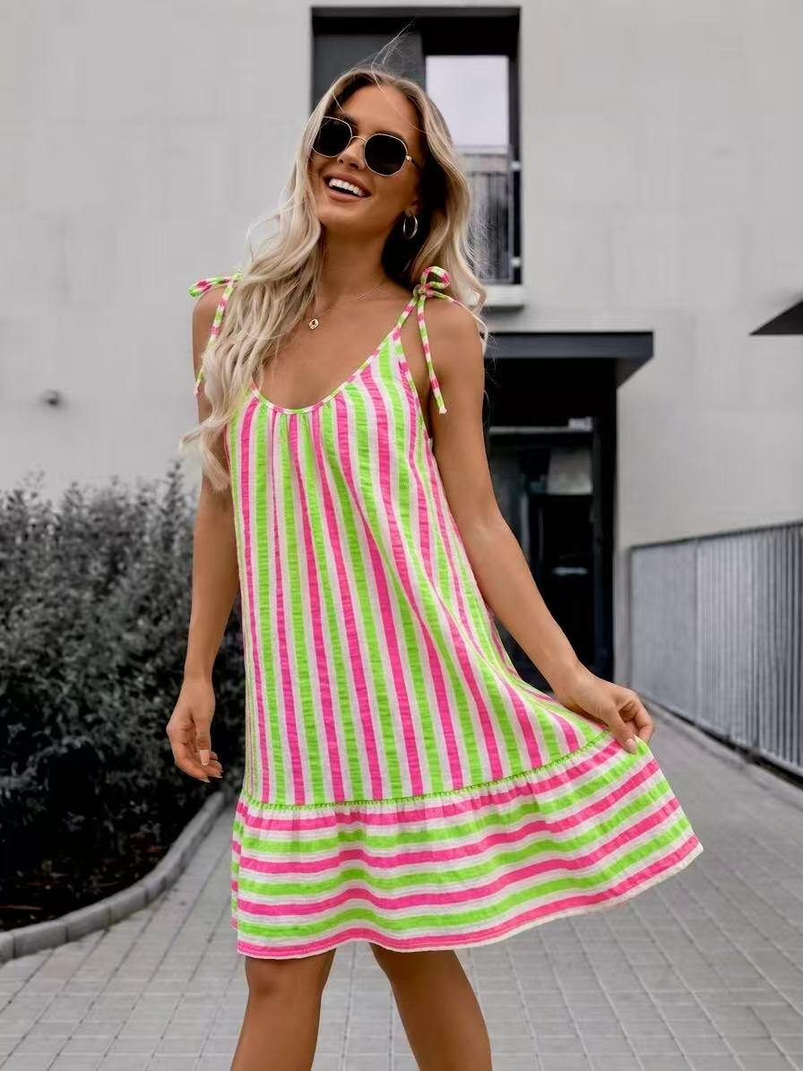 A woman wearing a sleeveless, tiered A-line dress with vertical pink and lime green stripes, styled with round sunglasses, standing outdoors on a paved walkway.