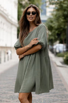 A woman wearing a textured sage green short-sleeve dress and black sunglasses, standing on a cobblestone street.