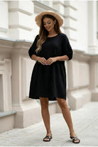 A woman wearing a black three-quarter sleeve shift dress, a beige straw hat, and black sandals, standing on a paved street in front of a white building.